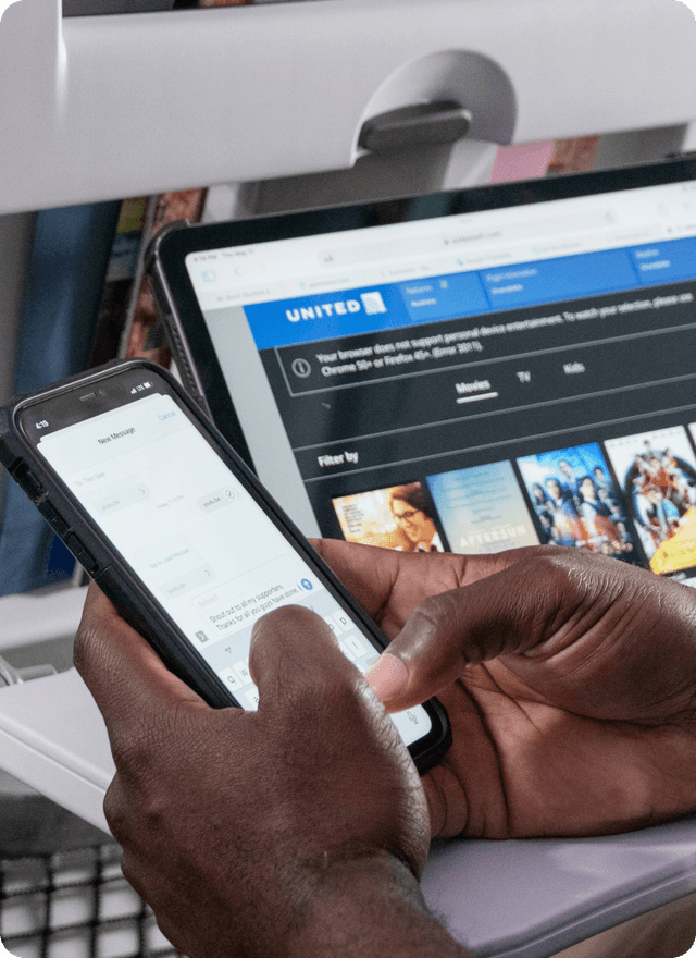 A passenger using a smartphone on an airplane with a United Airlines seatback entertainment screen visible in the background.