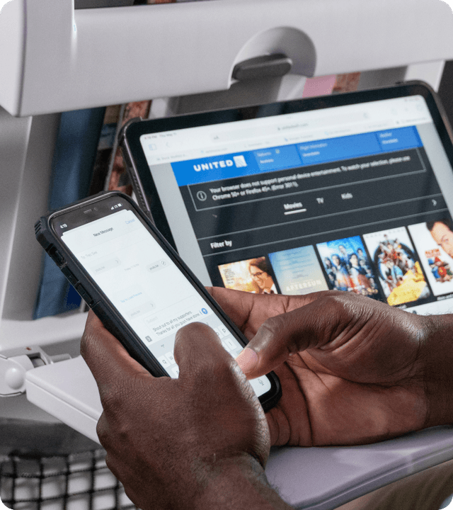 A passenger using a smartphone on an airplane with a United Airlines seatback entertainment screen visible in the background.
