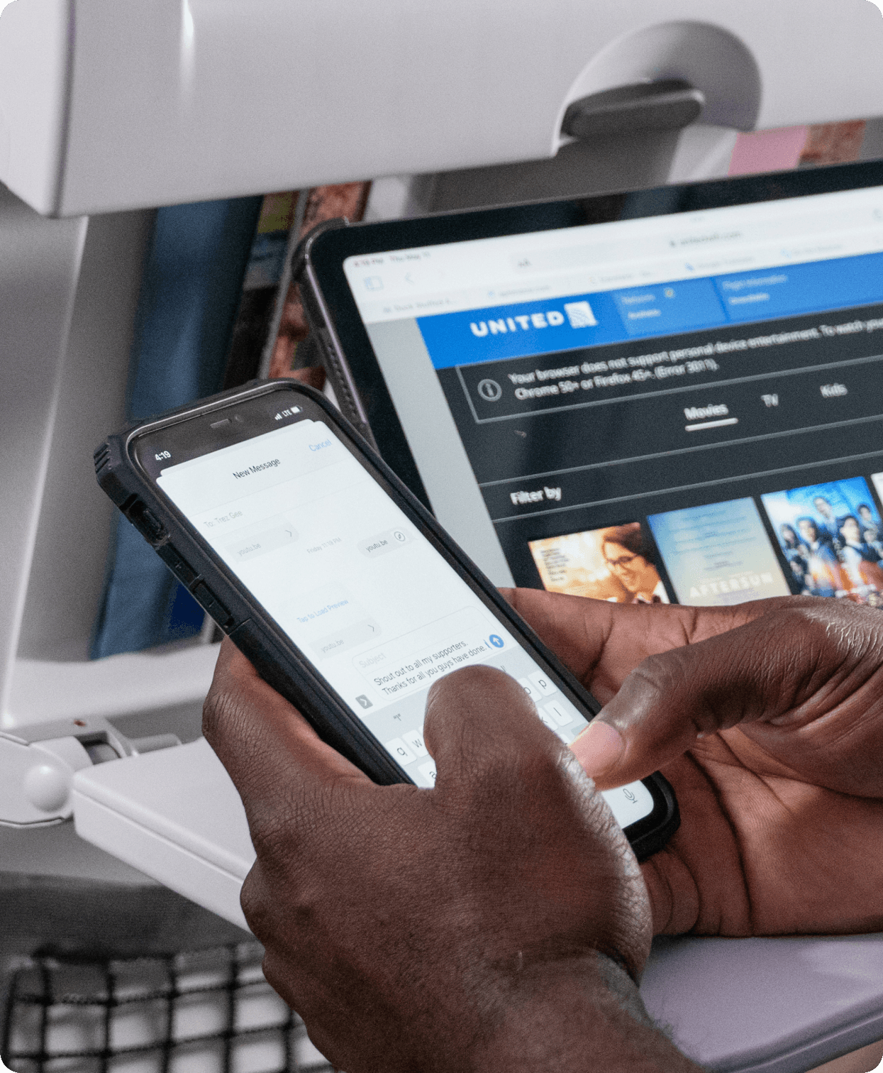 A passenger using a smartphone on an airplane with a United Airlines seatback entertainment screen visible in the background.