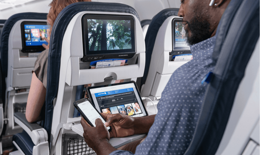 A man in an aisle seat looking at his cellphone with a laptop sitting on the tray table and a decorative image on the seat back display.