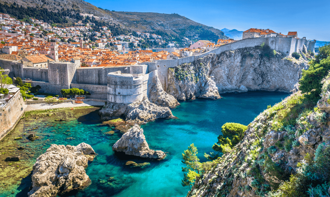 A long shot of an inlet with crystal clear water flanked by a rocky coastline with a rock wall sitting on top which surrounds an old city.