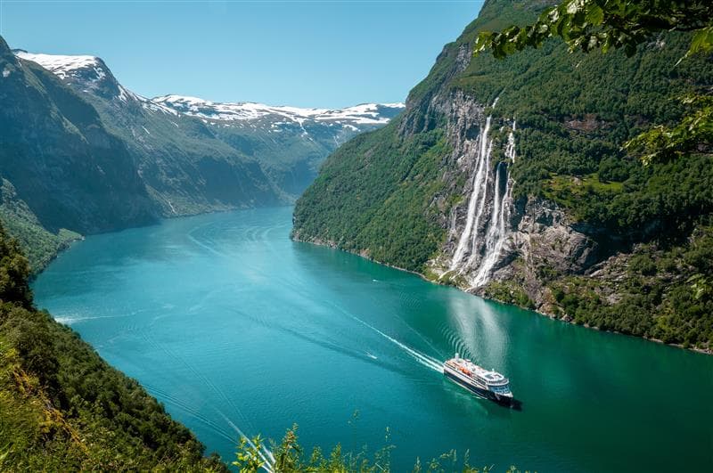 A distant shot of a cruise liner in a wide river gorge, flanked by majestic, snow capped, mountains on each side.