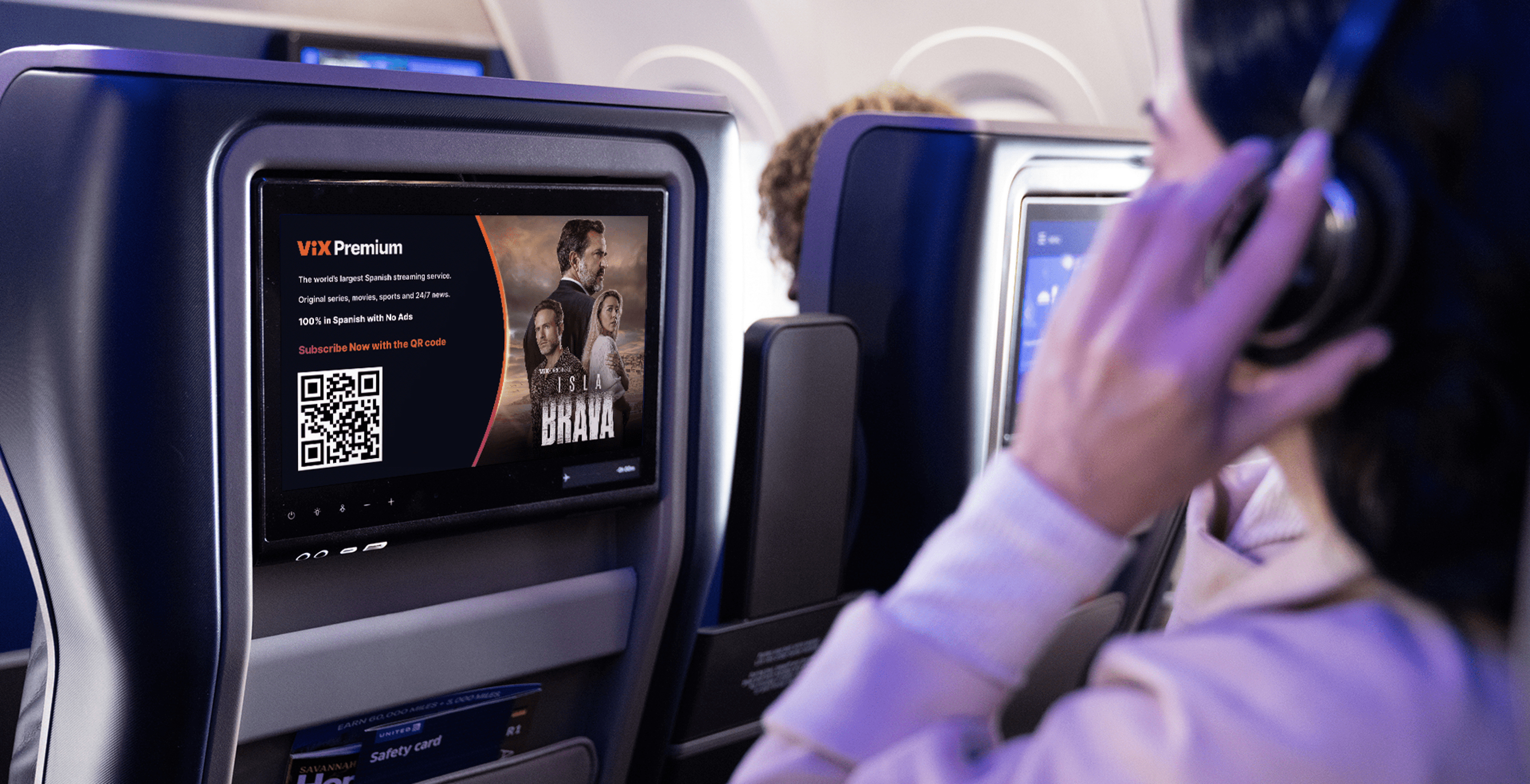 Woman sitting on a plane, watching the seat back monitor while she touches her wireless headphones.