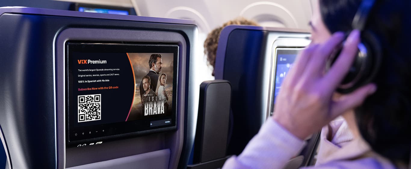 Woman sitting on a plane, watching the seat back monitor while she touches her wireless headphones.