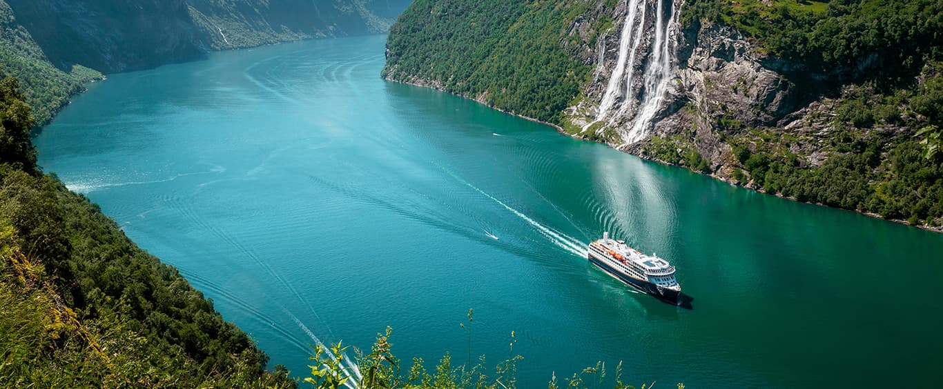 A distant shot of a cruise liner in a wide river gorge, flanked by majestic, snow capped, mountains on each side.