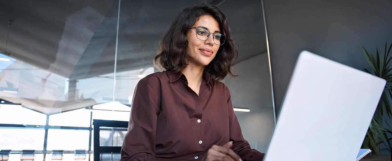 A blue hued moody, tightly cropped, photo of a woman typing on a laptop keyboard with her cell phone facing up by its side