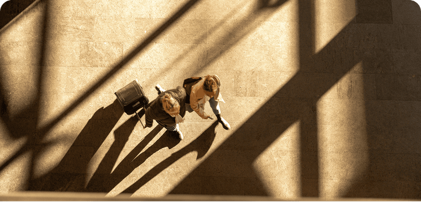 Two travelers walking with rolling suitcases across a sunlit floor with dramatic shadow patterns.