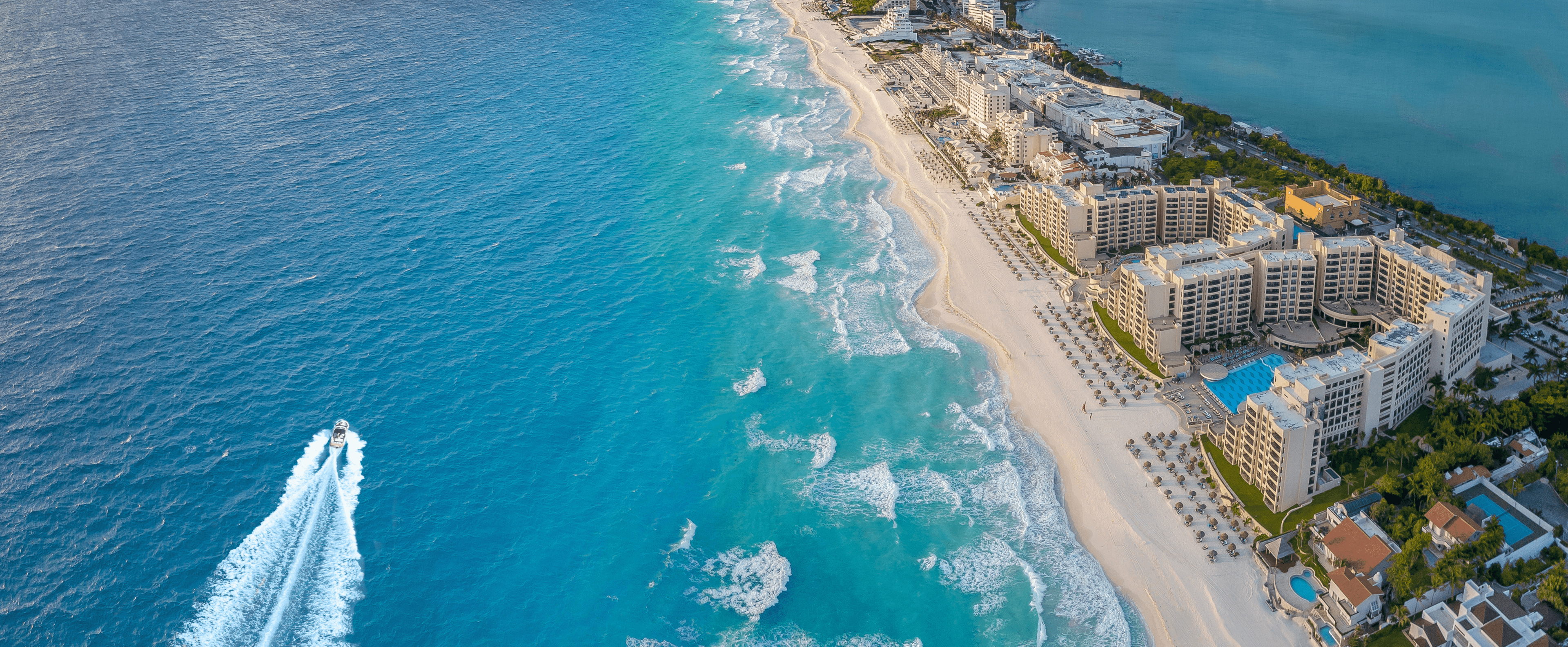 An aerial view of a long strip of beach with hotels and vacation houses flanked by ocean on both sides.  A speed boat is seen on the bottom left of the image