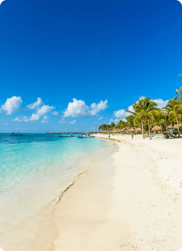 A photo of a beach with clear blue water and some small fishing boats. There are small huts and palm trees where people are relaxing and having fun.