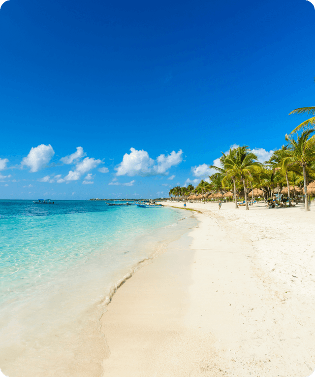 A photo of a beach with clear blue water and some small fishing boats. There are small huts and palm trees where people are relaxing and having fun.