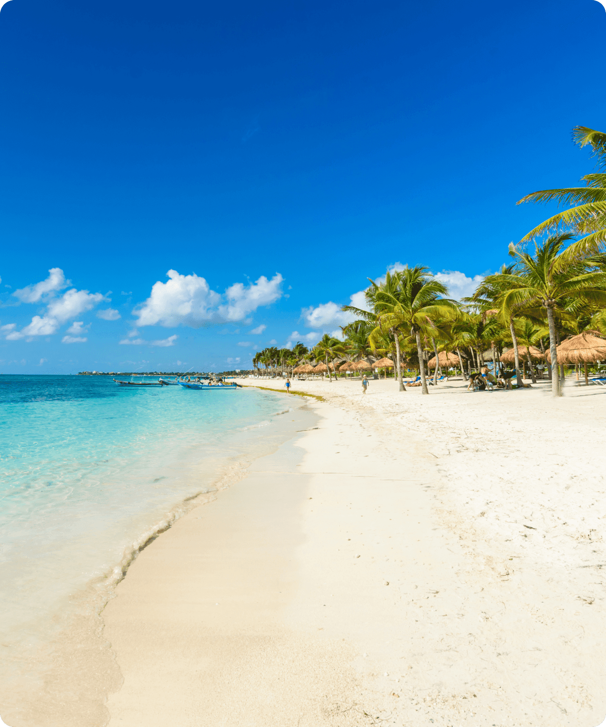A photo of a beach with clear blue water and some small fishing boats. There are small huts and palm trees where people are relaxing and having fun.