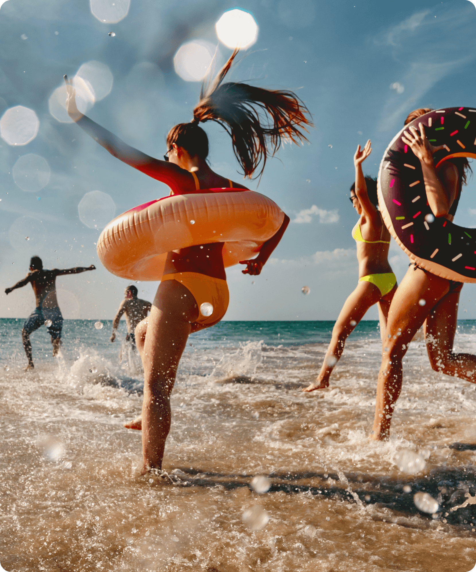 Group of friends playing in ocean waves with inflatable rings on a sunny beach day.