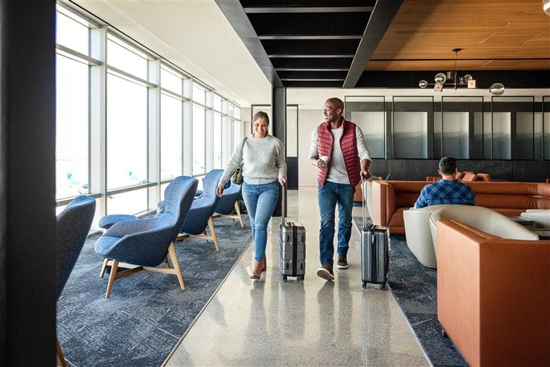 A smiling male and female traveler, walking through an airport lounge with roller suitcases.