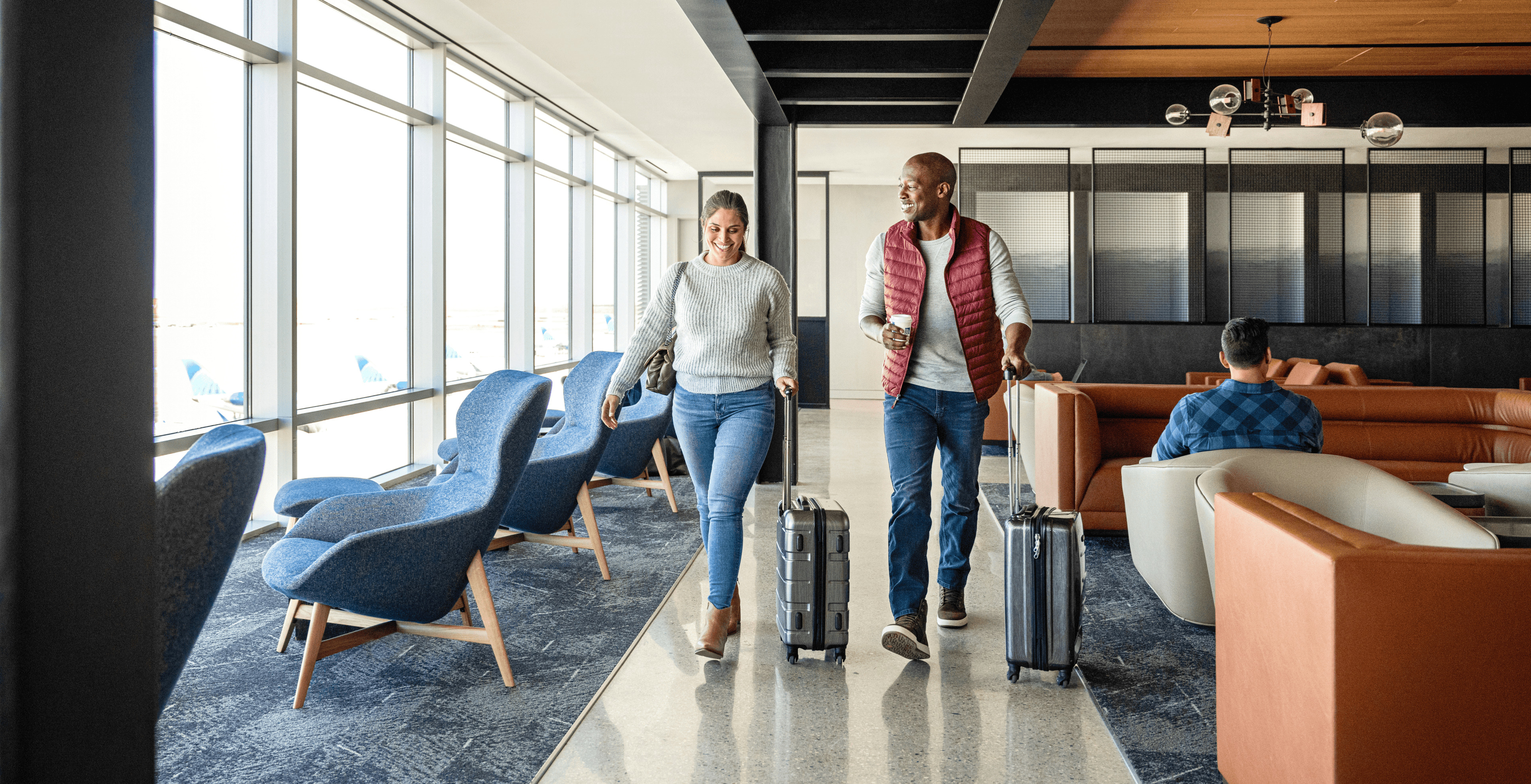 A smiling male and female traveler, walking through an airport lounge with roller suitcases.