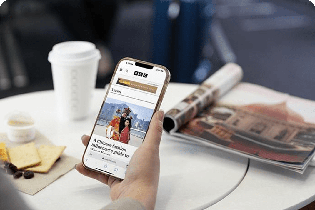 Person holding a smartphone while planning a trip with coffee and magazines nearby.