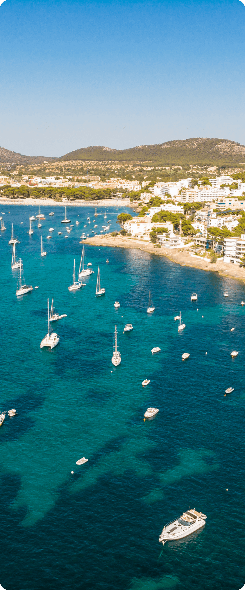 Aerial view of a coastal town with white buildings along the shore and numerous boats anchored in turquoise blue waters