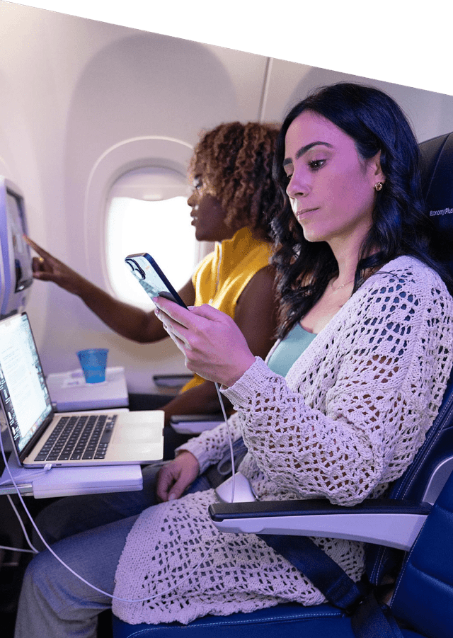 Woman on plane looking at her phone and laptop while woman next to her uses inflight entertainment screen.