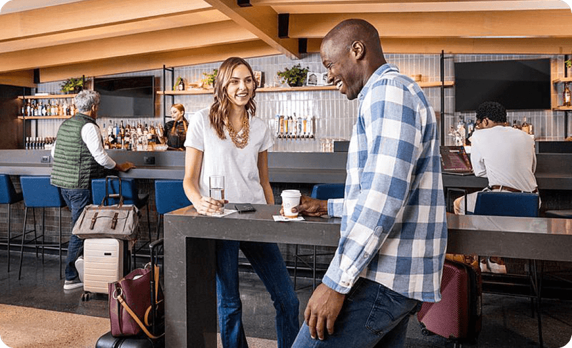Two travelers having a conversation at a table in an airport lounge with other passengers in the background