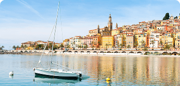 Colorful seaside town with a sailboat floating on calm reflective water under a clear blue sky.