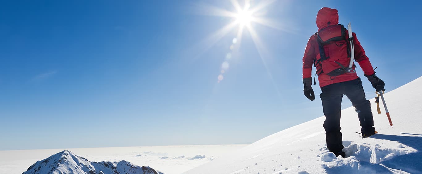 A mountain climber with a red jacket and climbing gear standing atop a snow covered mountain with a distant snow covered mountain peak to his left