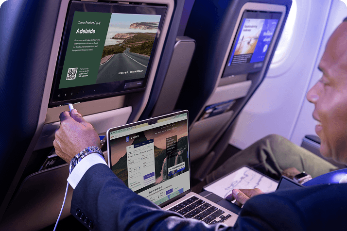 A man in an aisle seat looking at his cellphone with a laptop sitting on the tray table and a decorative image on the seat back display.